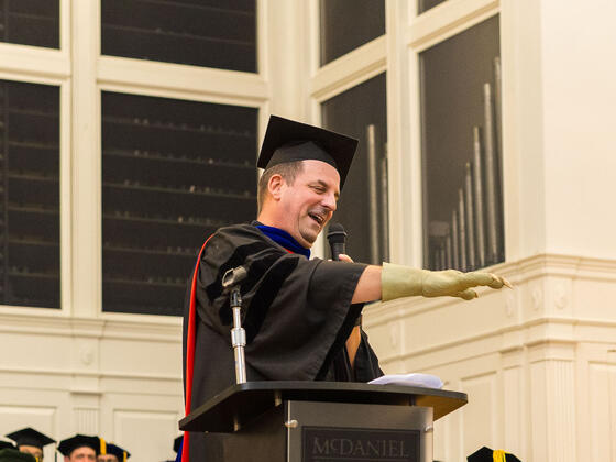 Faculty Member dressed in regalia speaks at podium while holding out arm with a Star Wars themed glove on his hand.