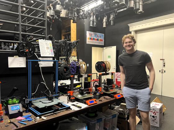 A student in a T-shirt and shorts stands next to a table of 3D print machines.