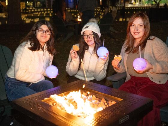 Three students hold light-up cups while eating s'mores by a firepit.
