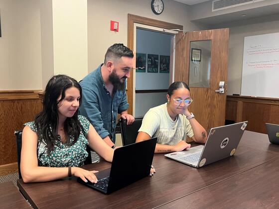 Two female students sit in front of laptops at a table and lecturer Alexander Champoux-Crowley looks down at one of their screens.