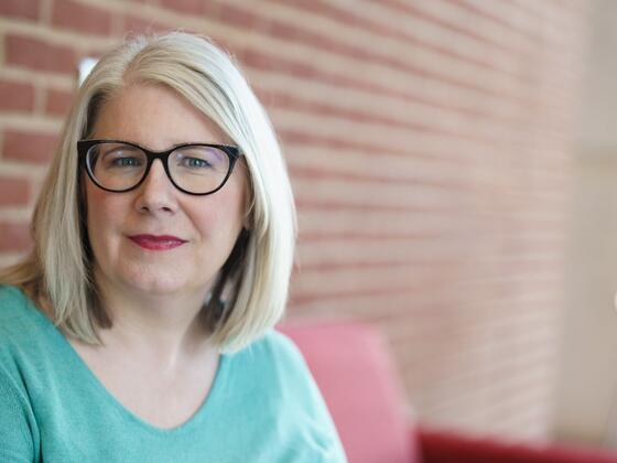 Headshot of Professor Stephanie Madsen with glasses and a blue shirt next to a brick building