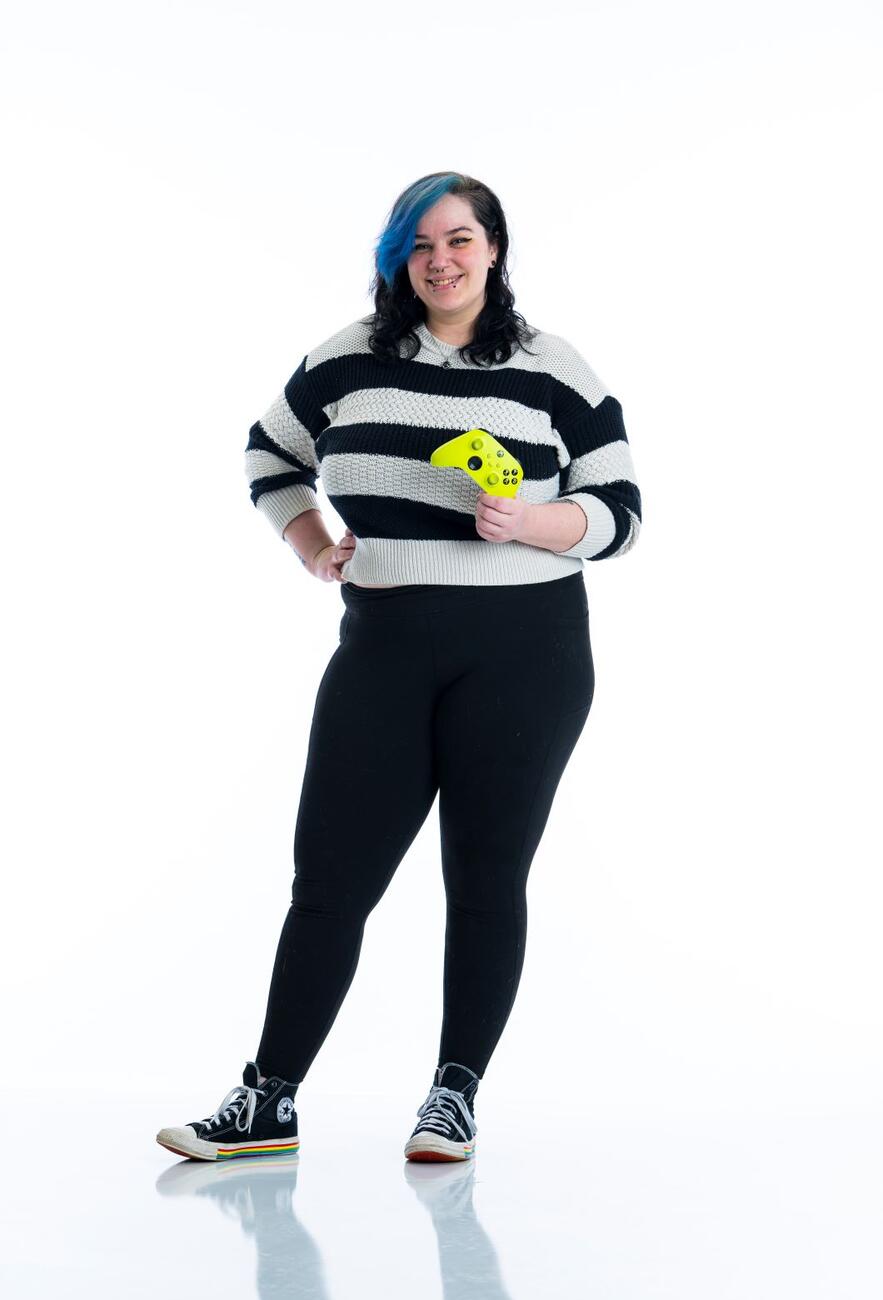 A student in a striped sweater stands with a hand on their hip while holding a green game controller.