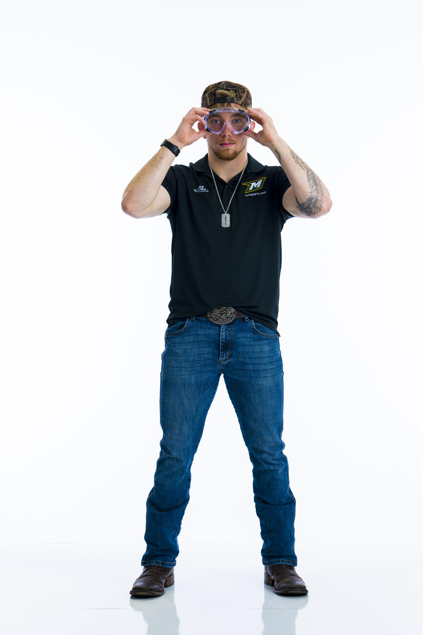 A student stands with his hands up to a pair of lab goggles he is wearing in front of a white background.