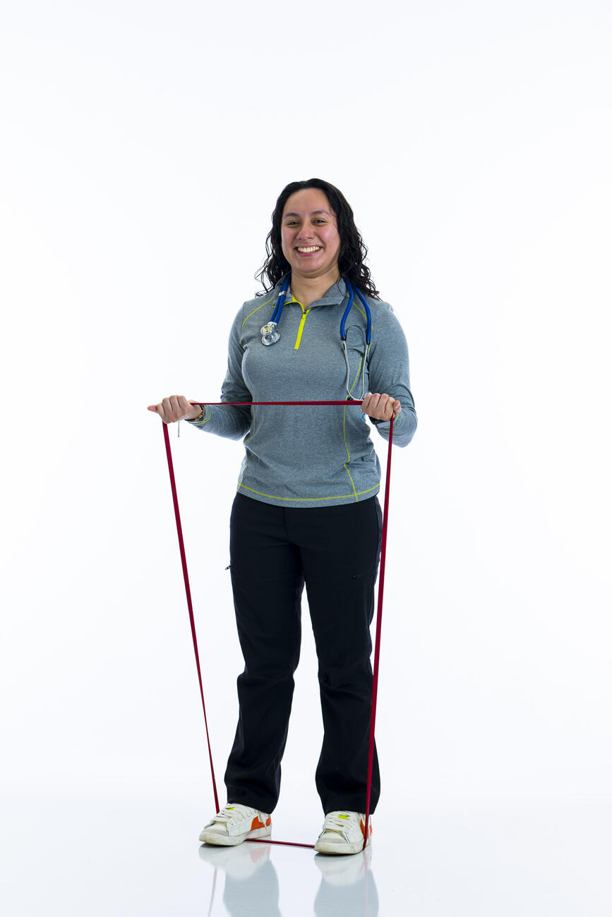 A student stands on a resistance band while stretching it in her hands while standing in front of white background.