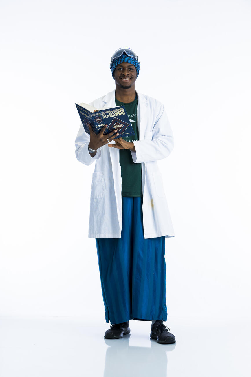 A student in a lab coat hold an Arabic book open while smiling at camera in front of a white background.