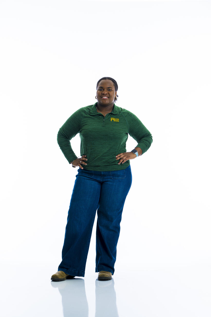 A student in a dark green shirt and jeans stands with hands on hips in front of a white background.