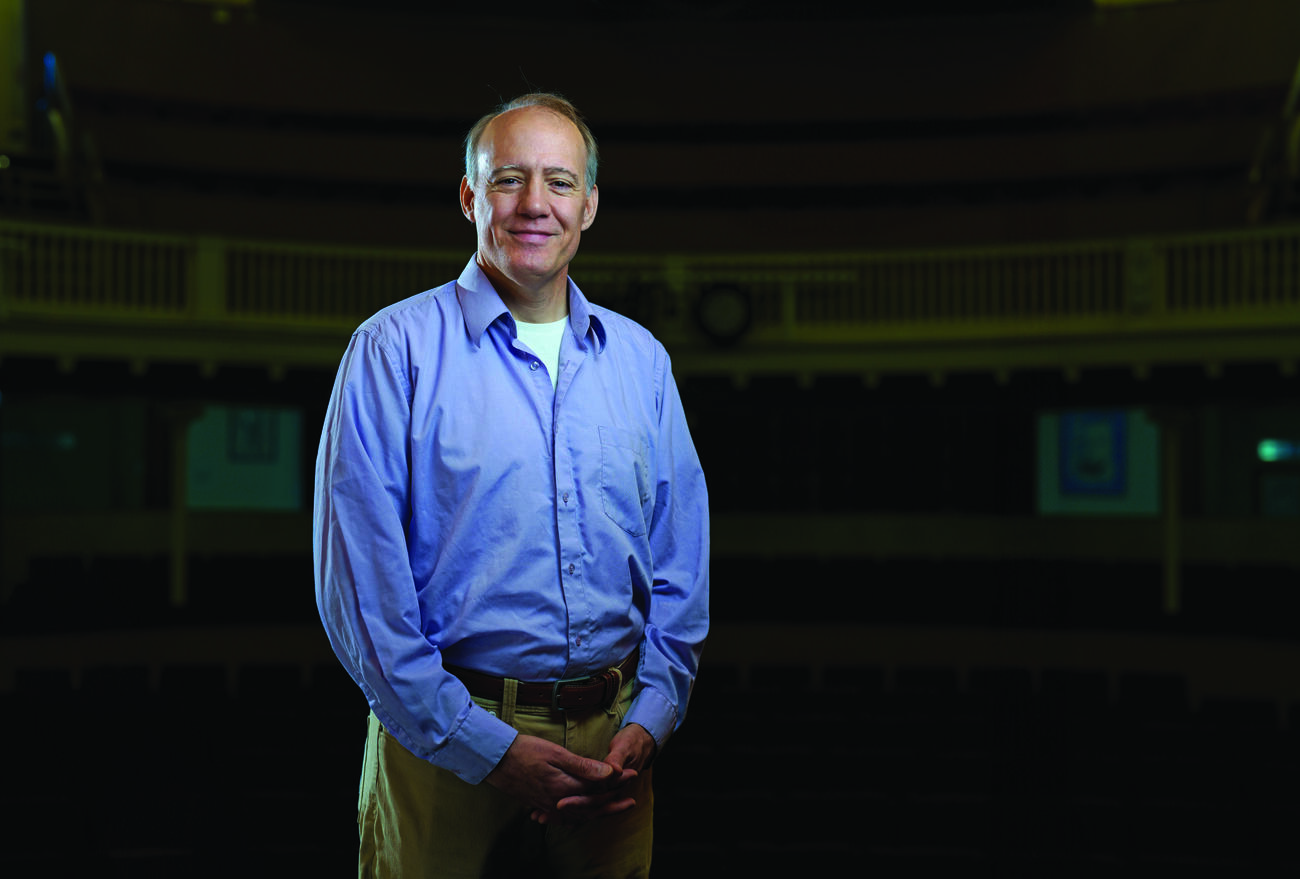 Professor Richard Sautter stands in a blue button up in the campus theatre.