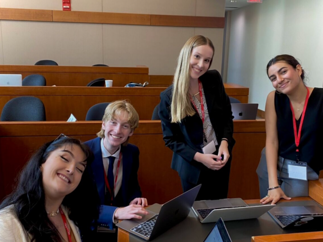 Photo of three female and one male wearing suits and lanyards gathered around a laptop while smiling.