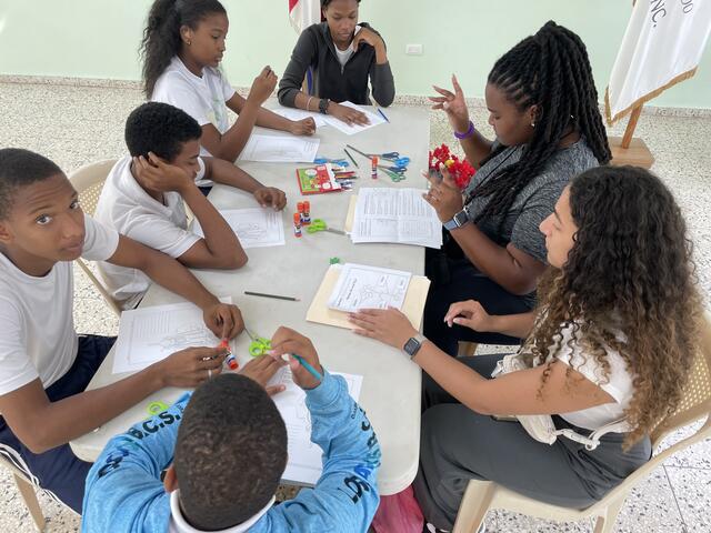 Two women sit at a table with five children in school uniforms and show them how to make ASL signs.