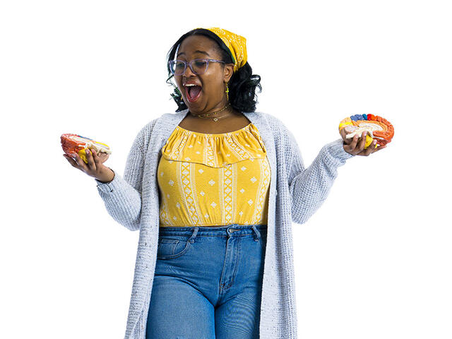 A student in a yellow shirt and jeans holds her hands out to either side while holding two halves of a model brain.