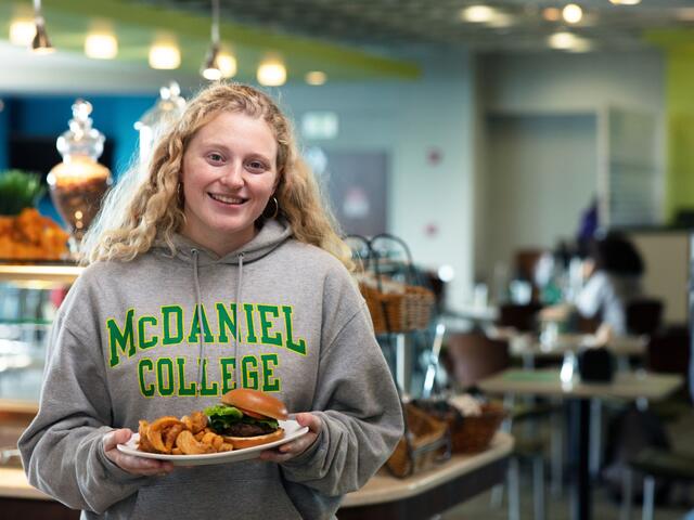 A female student in a McDaniel College sweatshirt stands in the dining hall holding a plate with a burger on it.