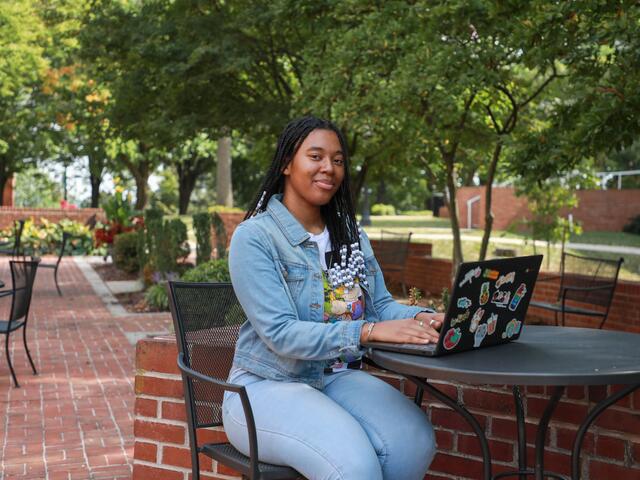 A student wearing a jean jacket sits at an outdoor table with a laptop in front of her.