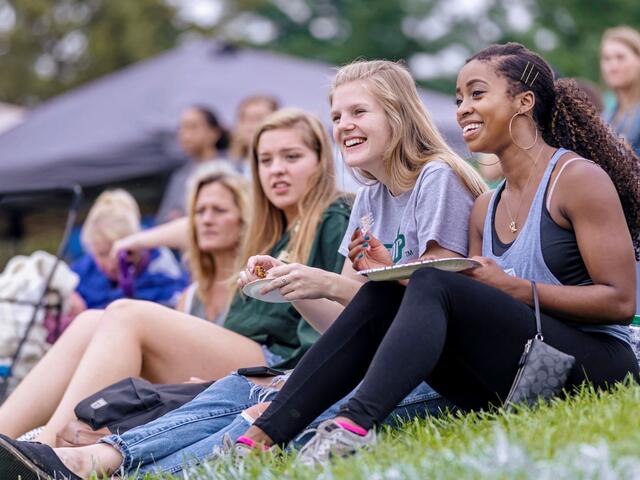 A group of four female students sit on the grass while holding plates and laughing.