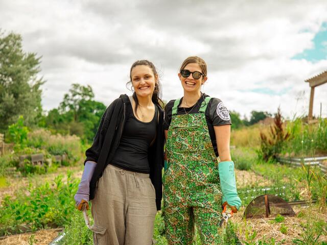 A student and professor pose outside in a garden while wearing gardening gloves.