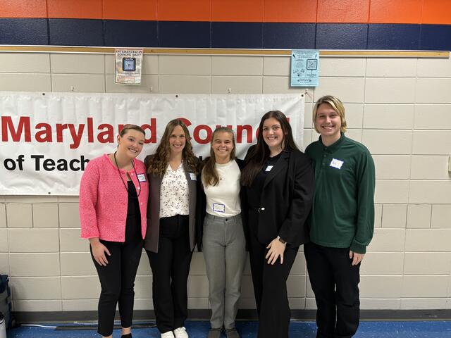 Four Education students and a faculty member pose in front of a banner that reads Maryland Council of Teachers of Mathematics.