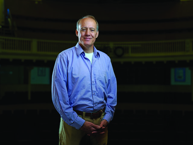 Professor Richard Sautter stands in a blue button up in the campus theatre.