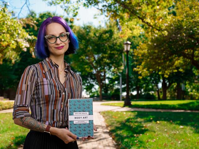 White woman holding a blue book with the title "Body Art" while standing outside.