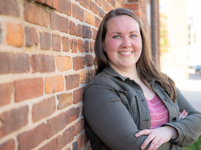 Photo of Robyn Forney standing in front of a brick wall, arms crossed, wearing a dark green button up and a pink shirt.