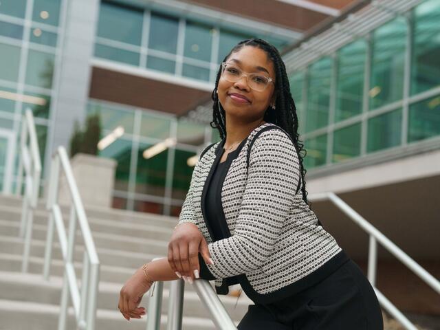Nakayla Lawson stands outside of the student center wearing a black shirt and pants with a black and white blazer. She leans forward on a railing.