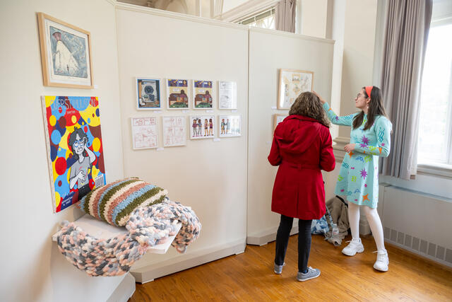 A student speaks with a visitor in an art gallery while gesturing to artworks on the walls.