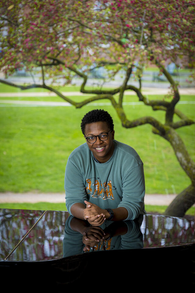 A student leans forward on a piano in front of a window with a tree behind him.