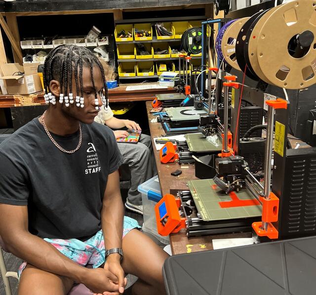 A student sits and watches a 3D printer.