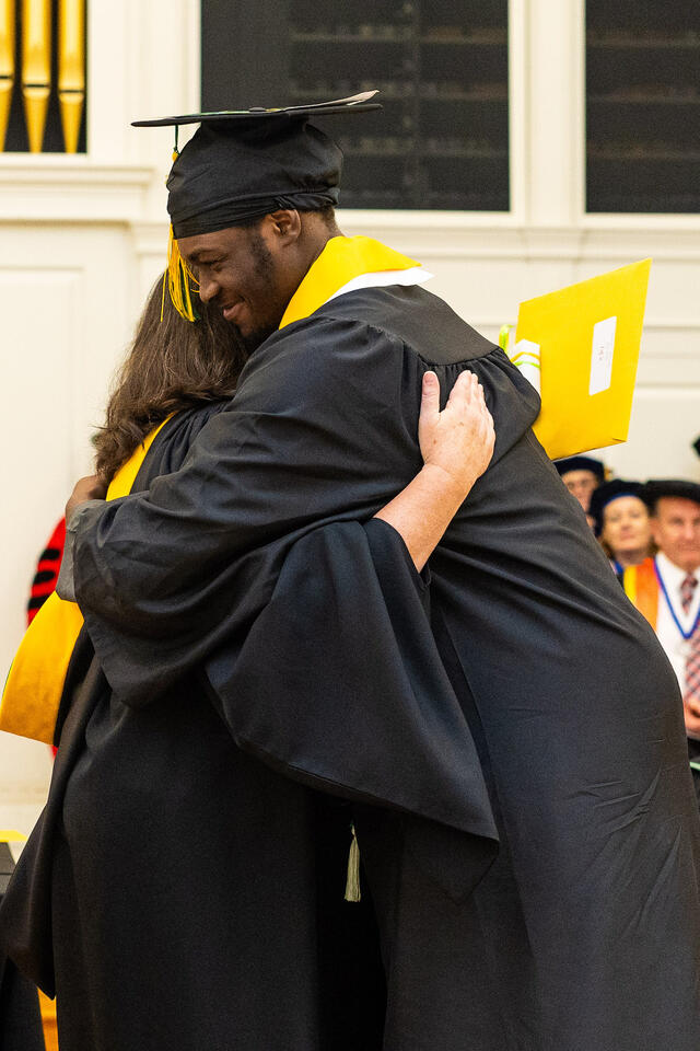 student in graduation regalia hugs faculty member