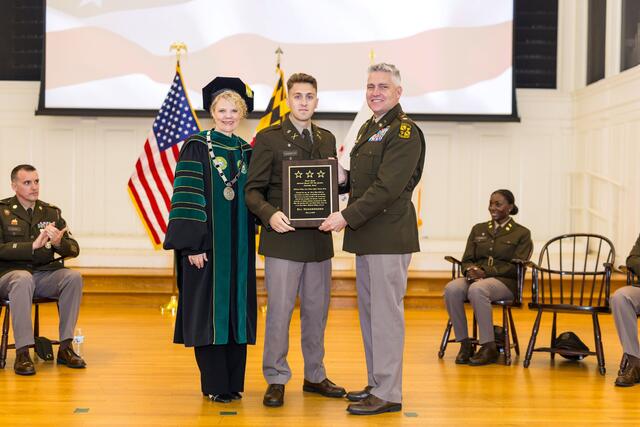 A cadet holds a plaque for the Lieutenant General (R) Otto Guenther Leadership Award while posing with college President Julia Jasken and Lieutenant Colonel Seth Hartmann.