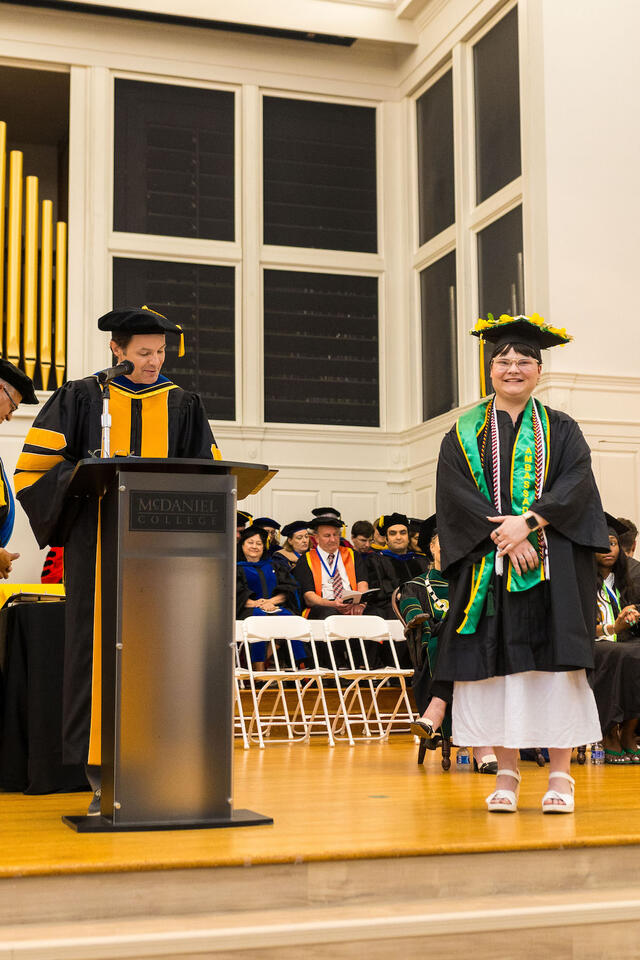 Student in graduation regalia poses while faculty member in regalia speaks at podium