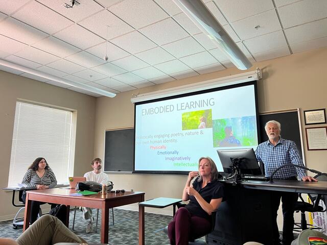 A professor stands at a podium in a classroom with two students seated at a table next to him. A screen behind him reads "Embodied Learning."