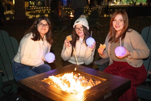Three students hold light-up cups while eating s'mores by a firepit.