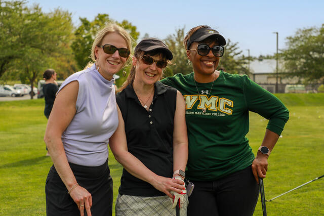Three women wearing athletic gear and sunglasses pose together while holding golf clubs.