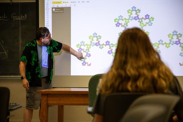 Photo taken from behind a seated student, looking at a McDaniel professor gesturing to a projection of a fractal.