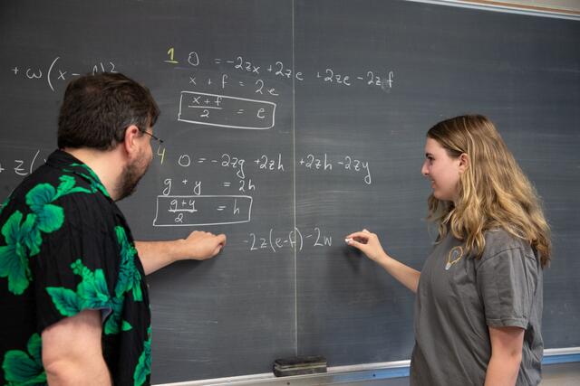 A student and professor write an equation on a chalkboard.