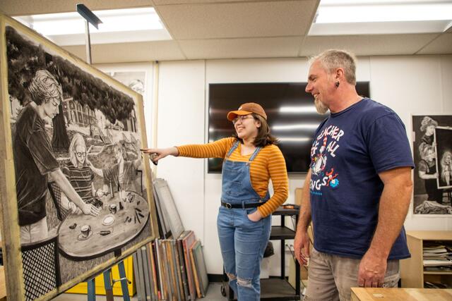 Professor Steve Pearson looks at artwork on an easel while a student in a ballcap and overalls points to it.