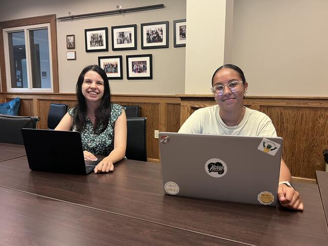 Two McDaniel research students smile at the camera while sitting with laptops in front of them.