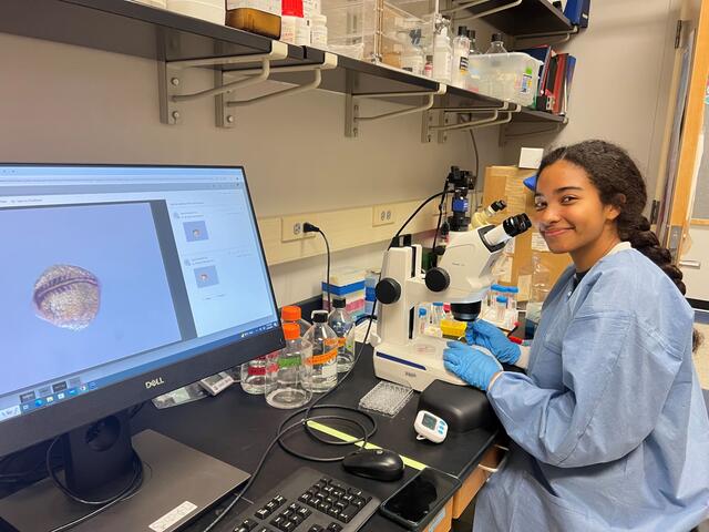 A student in a blue lab coat smiles at the camera while sitting in front of a microscope. An image of a zebrafish embryo from under the microscope appears on a computer screen next to her.