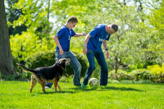 A dog runs next to a father and son playing soccer outside.