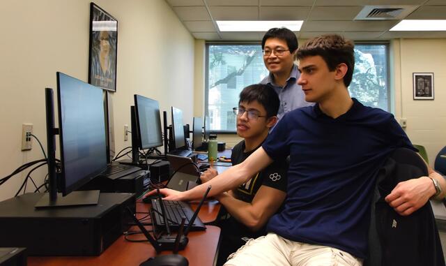 Two students looks at a computer screen while a faculty member looks over their shoulder.