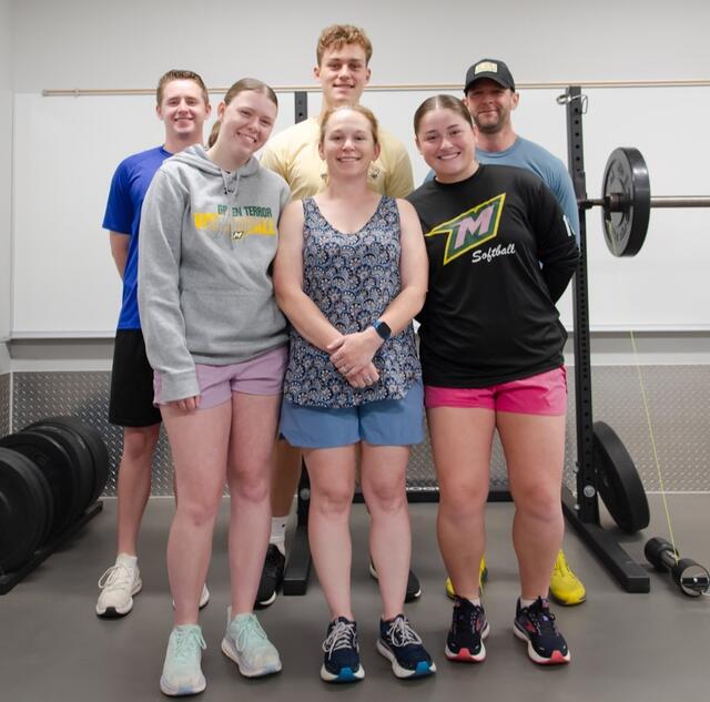 A group photo of two professors and four students standing next to a barbell rack.