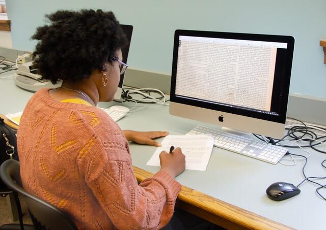 A student in an orange cardigan writes on a piece of paper while looking at an image of a newspaper story on a computer screen.