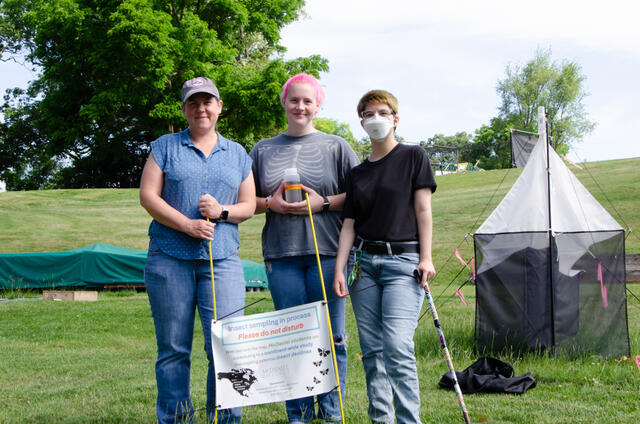 Two students pose with Professor Holly Martins outside next to a Malaise trap and behind a sign that reads "Insect sampling in process please do not disturb."