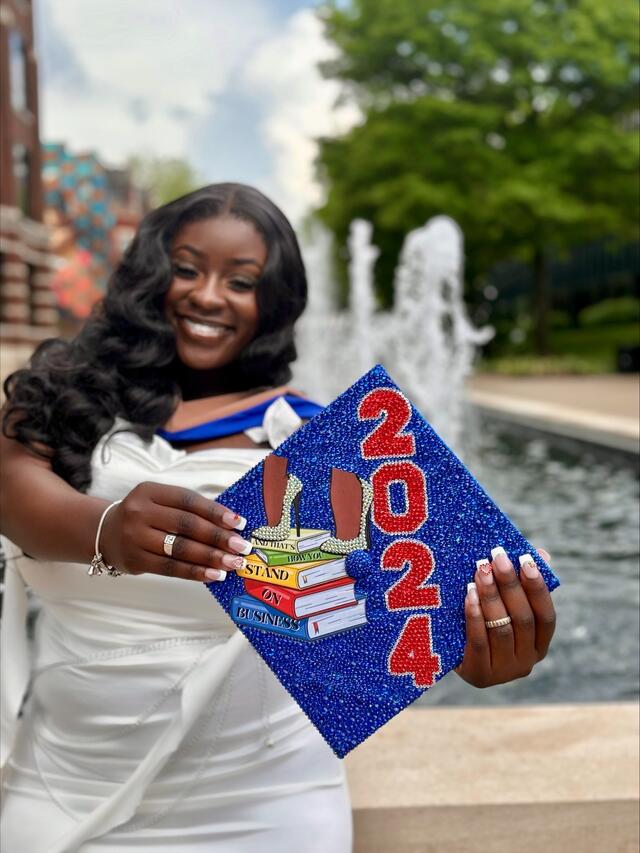 A Black woman in a white dress stands in front of a fountain and holds out a graduation mortar board that reads "2024" with an image of two high heels on a stack of books.