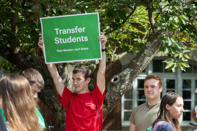 student wearing red shirt holding up sign that says "Transfer Students"