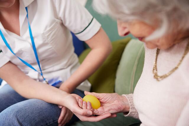 A close up image of a woman's hands helping an older woman hold a yellow egg.