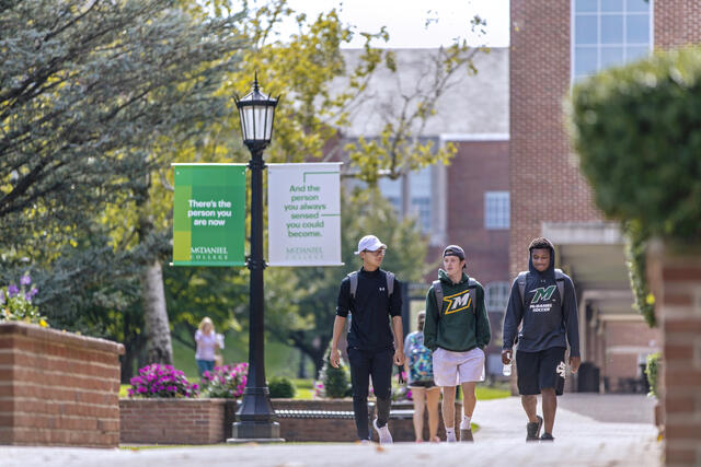 3 studetns walk on campus next to green trees and lamppost banners