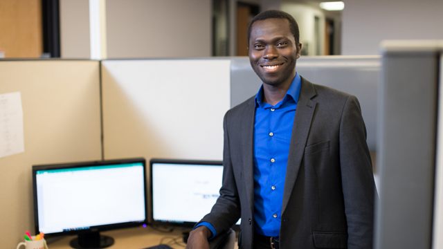 Data Analytics M.S. student Michael Eniolade wears a suit and stands next to a computer screen on a desk.