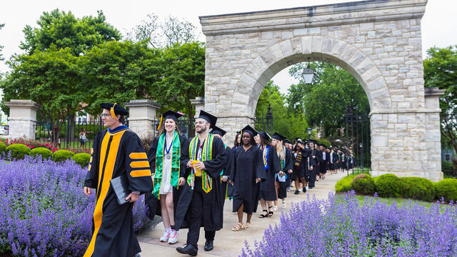 Students in regalia walk through the historical arch, led by the provost in regalia