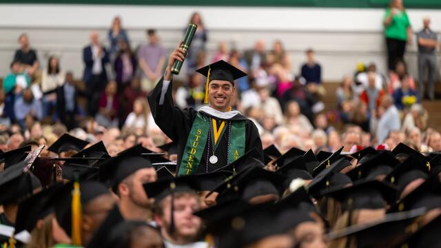 A student in a graduation cap and gown holds up his degree while standing in a crowd of seated graduates.