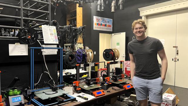 A student in a T-shirt and shorts stands next to a table of 3D print machines.